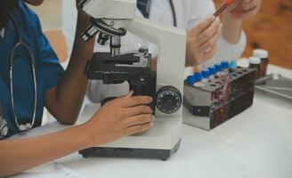 Medical worker in lab coat and sterile mask, doing a microscope analysis while her colleague are working behind photo