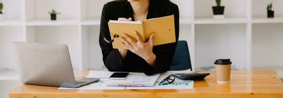 mujer de negocios asiática que usa una computadora portátil y trabaja en la oficina con un documento de calculadora en el escritorio, planificando el análisis del informe financiero, la inversión del plan de negocios, el concepto de análisis financiero. foto