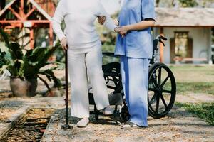 Asian young woman nurse at nursing home take care disabled senior man. Caregiver doctor serve physical therapy for older elderly patient to exercise and practice walking on walker or cane at backyard. photo