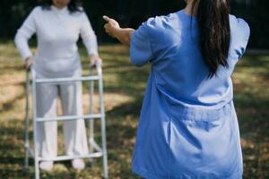 asiático joven mujer enfermero a enfermería hogar tomar cuidado discapacitado mayor hombre. cuidador médico servir físico terapia para más viejo mayor paciente a ejercicio y práctica caminando en caminante o caña a patio interior. foto