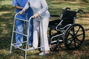 Asian young woman nurse at nursing home take care disabled senior man. Caregiver doctor serve physical therapy for older elderly patient to exercise and practice walking on walker or cane at backyard. photo