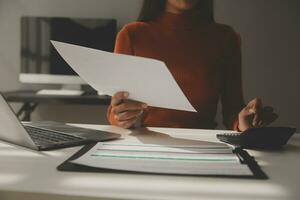 mujer de negocios asiática que usa una computadora portátil y trabaja en la oficina con un documento de calculadora en el escritorio, planificando el análisis del informe financiero, la inversión del plan de negocios, el concepto de análisis financiero. foto