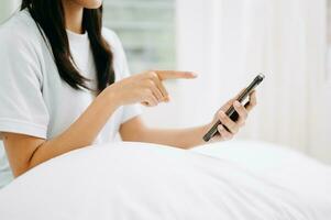 Beautiful young asian woman on a white bed with coffee cub and using smart phone at home. lifestyle concept photo