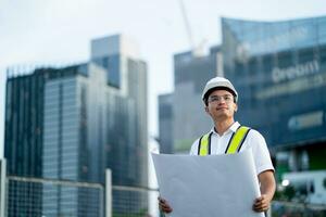 Asian male engineer holding blueprint and planning project Construction site background. Engineer planning a project and looking up at the sky. photo