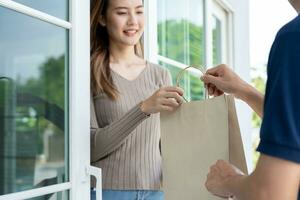 Happy smiling Asian woman receives paper bag parcel of food from courier front house. Delivery man send deliver express. online shopping, paper container, takeaway, postman, delivery service, packages photo