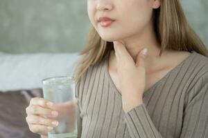 asiático mujer poner su mano en su cuello debido a un dolorido garganta. hembra amigdalitis causas un inflamación. tratado por tomando agua, tomando antibióticos, consultar un doctor, virus, corona virus, influenza, gripe foto