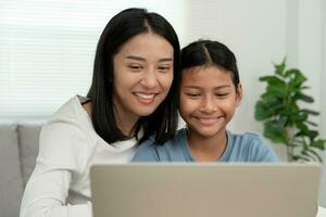 Mother teaching lesson for daughter by laptop. Asian young little girl learn at home. Do homework with kind mother help, encourage for exam. Asia girl happy Homeschool. Mom advise education together. photo