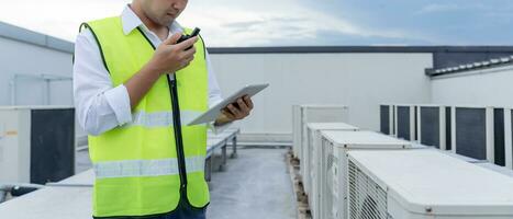 Asian maintenance engineer works on the roof of factory. contractor inspect compressor system and plans installation of air condition systems in construction. technology, online, mobile application. photo