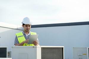 Asian maintenance engineer works on the roof of factory. contractor inspect compressor system and plans installation of air condition systems in construction. technology, online, mobile application. photo
