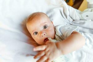 Cute little newborn girl drinking milk from bottle and looking at camera on white background. Infant baby sucking eating milk nutrition lying down on crib bed at home. Motherhood happy child concept. photo