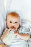 Cute little newborn girl drinking milk from bottle and looking at camera on white background. Infant baby sucking eating milk nutrition lying down on crib bed at home. Motherhood happy child concept. photo