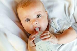 Cute little newborn girl drinking milk from bottle and looking at camera on white background. Infant baby sucking eating milk nutrition lying down on crib bed at home. Motherhood happy child concept. photo