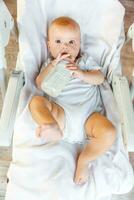 Cute little newborn girl drinking milk from bottle and looking at camera on white background. Infant baby sucking eating milk nutrition lying down on feeding chair at home. Motherhood happy child photo