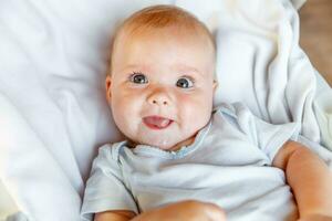 Cute little newborn girl with smiling face looking at camera on white background. Infant baby resting playing lying down on crib bed at home. Motherhood happy child concept. photo