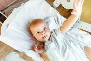 Cute little newborn girl drinking milk from bottle and looking at camera on white background. Infant baby sucking eating milk nutrition lying down on crib bed at home. Motherhood happy child concept. photo