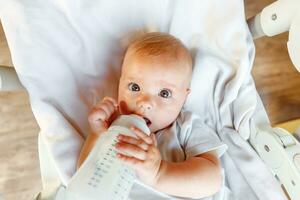 Cute little newborn girl drinking milk from bottle and looking at camera on white background. Infant baby sucking eating milk nutrition lying down on crib bed at home. Motherhood happy child concept. photo