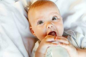 linda pequeño recién nacido niña Bebiendo Leche desde botella y mirando a cámara en blanco antecedentes. infantil bebé succión comiendo Leche nutrición acostado abajo en cuna cama a hogar. maternidad contento niño concepto. foto