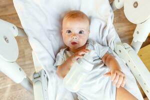 linda pequeño recién nacido niña Bebiendo Leche desde botella y mirando a cámara en blanco antecedentes. infantil bebé succión comiendo Leche nutrición acostado abajo en alimentación silla a hogar. maternidad contento niño foto