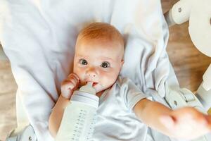 Cute little newborn girl drinking milk from bottle and looking at camera on white background. Infant baby sucking eating milk nutrition lying down on feeding chair at home. Motherhood happy child photo