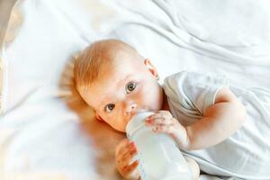 Cute little newborn girl drinking milk from bottle and looking at camera on white background. Infant baby sucking eating milk nutrition lying down on crib bed at home. Motherhood happy child concept. photo