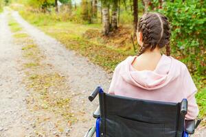 joven discapacitada feliz en silla de ruedas en la carretera en el parque del hospital esperando los servicios del paciente. niña paralizada en silla inválida para personas discapacitadas al aire libre en la naturaleza. concepto de rehabilitación. foto