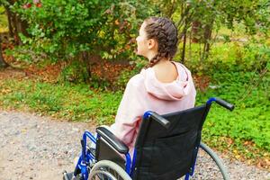 Young happy handicap woman in wheelchair on road in hospital park waiting for patient services. Paralyzed girl in invalid chair for disabled people outdoor in nature. Rehabilitation concept. photo