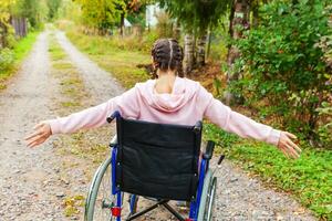 Young happy handicap woman in wheelchair on road in hospital park enjoying freedom. Paralyzed girl in invalid chair for disabled people outdoor in nature. Rehabilitation concept. photo