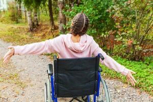 Young happy handicap woman in wheelchair on road in hospital park enjoying freedom. Paralyzed girl in invalid chair for disabled people outdoor in nature. Rehabilitation concept. photo