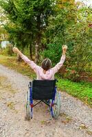 Young happy handicap woman in wheelchair on road in hospital park enjoying freedom. Paralyzed girl in invalid chair for disabled people outdoor in nature. Rehabilitation concept. photo