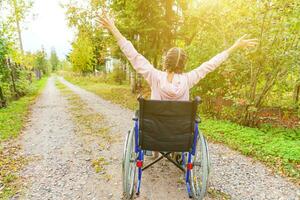 Young happy handicap woman in wheelchair on road in hospital park enjoying freedom. Paralyzed girl in invalid chair for disabled people outdoor in nature. Rehabilitation concept. photo