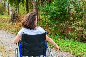 Young happy handicap woman in wheelchair on road in hospital park enjoying freedom. Paralyzed girl in invalid chair for disabled people outdoor in nature. Rehabilitation concept. photo