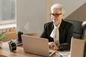 Confident stylish european middle aged senior woman using laptop at workplace. Stylish older mature 60s gray haired lady businesswoman sitting at office table. Boss leader teacher professional worker. photo