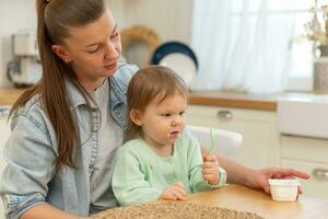 Happy family at home. Mother feeding her baby girl from spoon in kitchen. Little toddler child with messy funny face eats healthy food at home. Young woman mom giving food to kid daughter. photo