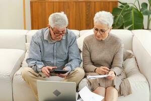 Middle aged senior couple sit with laptop and paper document. Older mature man woman reading paper bill pay online at home managing bank finances calculating taxes planning loan debt pension payment. photo
