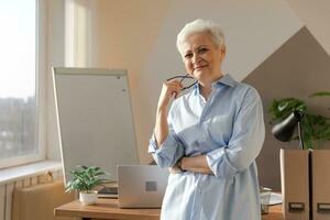 Portrait of confident stylish european middle aged senior woman at workplace. Stylish older mature 60s gray haired lady businesswoman in modern office. Boss leader teacher professional worker. photo