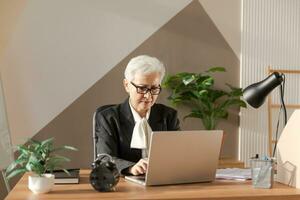 Confident stylish european middle aged senior woman using laptop at workplace. Stylish older mature 60s gray haired lady businesswoman sitting at office table. Boss leader teacher professional worker. photo