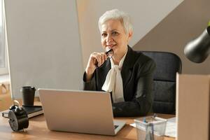 Portrait of confident stylish european middle aged senior woman at workplace. Stylish older mature 60s gray haired lady businesswoman sitting in modern office. Boss leader teacher professional worker. photo