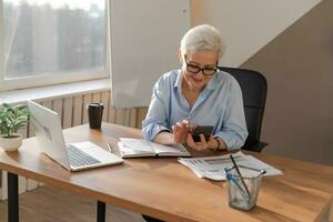 Confident stylish european middle aged senior woman using smartphone at workplace. Stylish older mature 60s gray haired lady businesswoman with cell phone in office. Boss leader using internet apps. photo