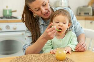 Happy family at home. Mother feeding her baby girl from spoon in kitchen. Little toddler child with messy funny face eats healthy food at home. Young woman mom giving food to kid daughter. photo