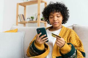 mujer afroamericana comprando en línea con un teléfono inteligente pagando con tarjeta de crédito dorada. niña sentada en casa comprando en internet ingrese los detalles de la tarjeta de crédito. servicio de entrega de comercio electrónico de compras en línea. foto