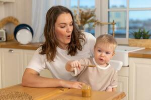 Happy family at home. Mother feeding baby in kitchen. Little boy with messy funny face eats healthy food. Child learns eat by himself holding spoon. Woman mom giving food to kid son. Self feeding. photo