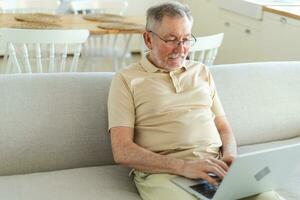 Confident stylish happy middle aged senior man using laptop at home. Stylish older mature 60s beard grandfather sitting at couch looking at computer screen typing chatting reading writing email. photo