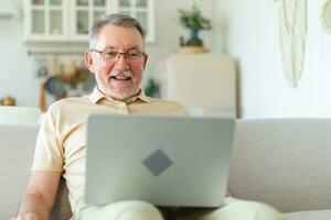 Confident stylish happy middle aged senior man using laptop at home. Stylish older mature 60s beard grandfather sitting at couch looking at computer screen typing chatting reading writing email. photo