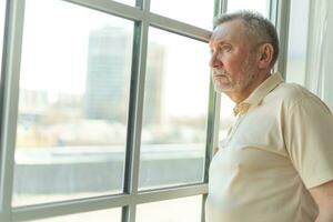 Sad tired lonely disappointed older senior man at home alone. Unhappy grandfather looking sadly outside window experiencing grief relative death bad news. Stressed elder man suffering from loneliness. photo