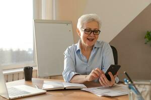 Confident stylish european middle aged senior woman using smartphone at workplace. Stylish older mature 60s gray haired lady businesswoman with cell phone in office. Boss leader using internet apps. photo