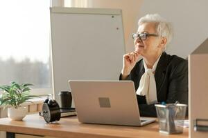 Confident stylish european middle aged senior woman using laptop at workplace. Stylish older mature 60s gray haired lady businesswoman sitting at office table. Boss leader teacher professional worker. photo