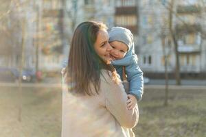 contento familia exterior. madre abrazando su niño exterior. mamá levantamiento en aire pequeño niñito niño hijo. mujer y pequeño bebé chico descansando caminando en parque. madre abrazos bebé con amor cuidado. foto