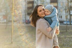 contento familia exterior. madre abrazando su niño exterior. mamá levantamiento en aire pequeño niñito niño hijo. mujer y pequeño bebé chico descansando caminando en parque. madre abrazos bebé con amor cuidado. foto