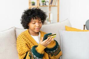mujer afroamericana comprando en línea con un teléfono inteligente pagando con tarjeta de crédito dorada. niña sentada en casa comprando en internet ingrese los detalles de la tarjeta de crédito. servicio de entrega de comercio electrónico de compras en línea. foto