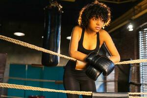 Woman fighter girl power. African american woman fighter with boxing gloves standing on boxing ring leaning on ropes waiting and resting. Strong powerful girl. Strength fit body workout training. photo